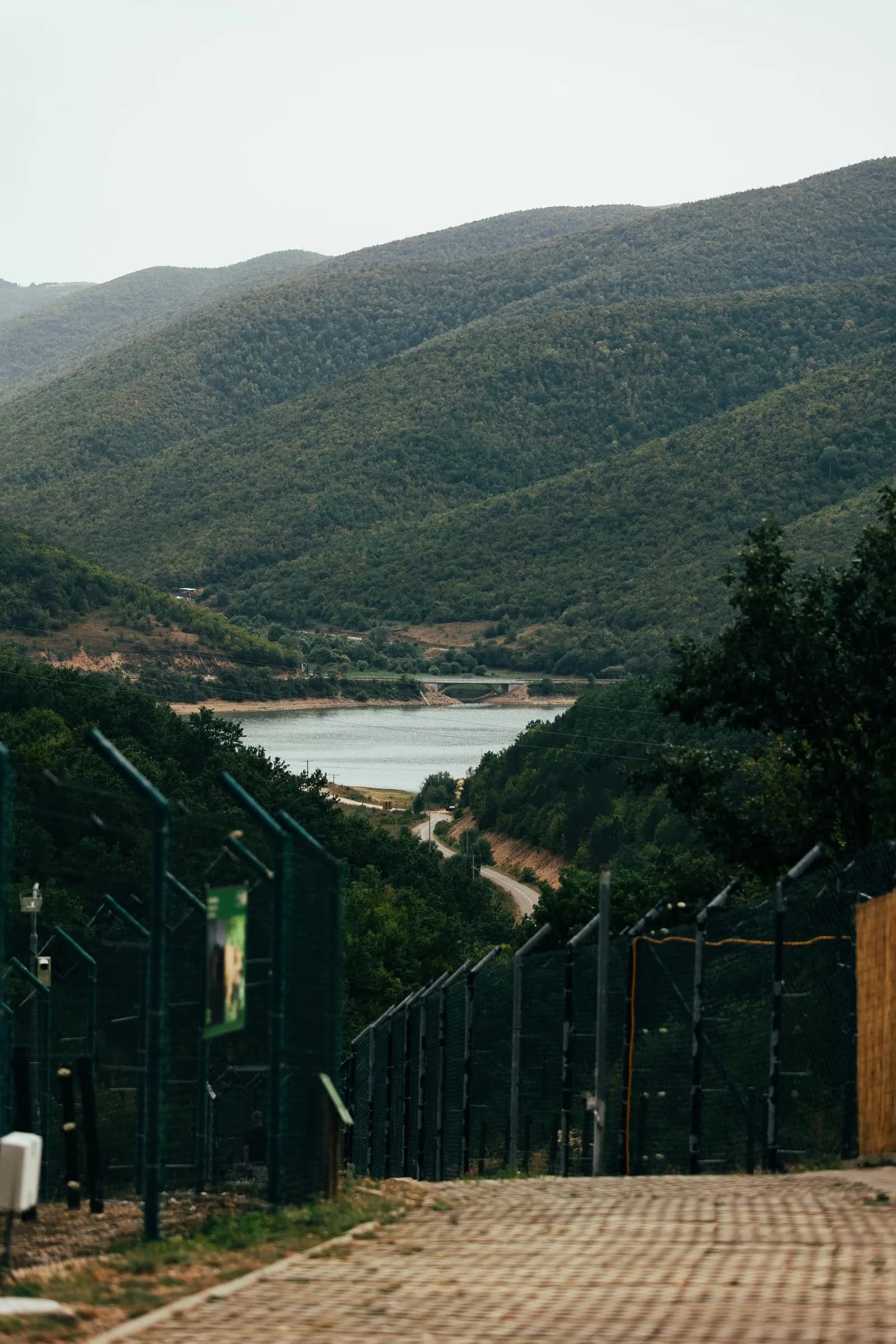 A forested valley and lake seen from the sanctuary path, beside a tall fence.